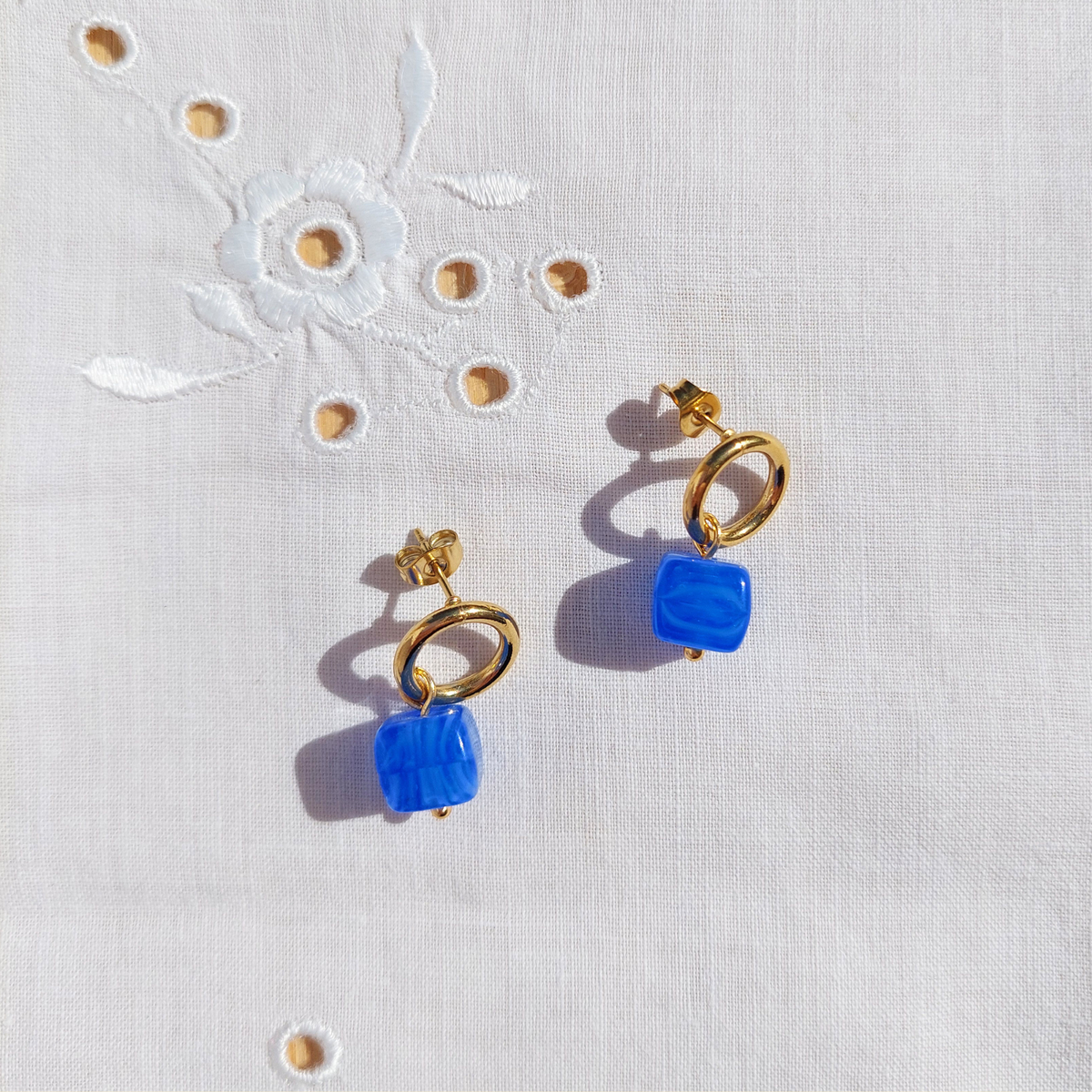 A pair of gold-tone drop earrings with square blue glass pendants, displayed on a white embroidered fabric background.