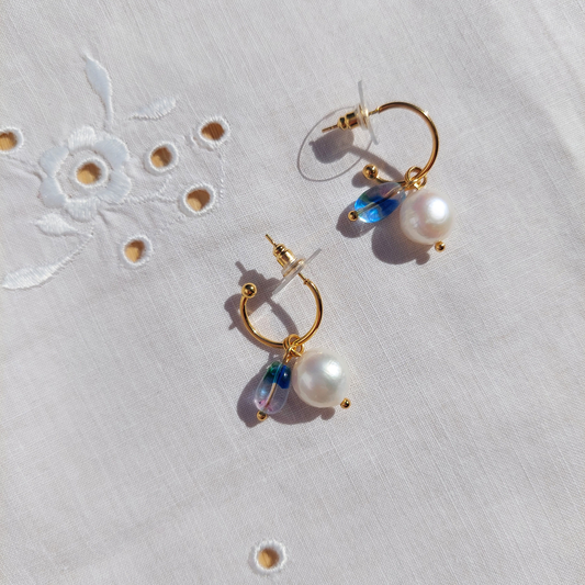 A pair of gold hoop earrings with dangling white pearls and iridescent blue-green teardrop beads, displayed on a white embroidered fabric background.