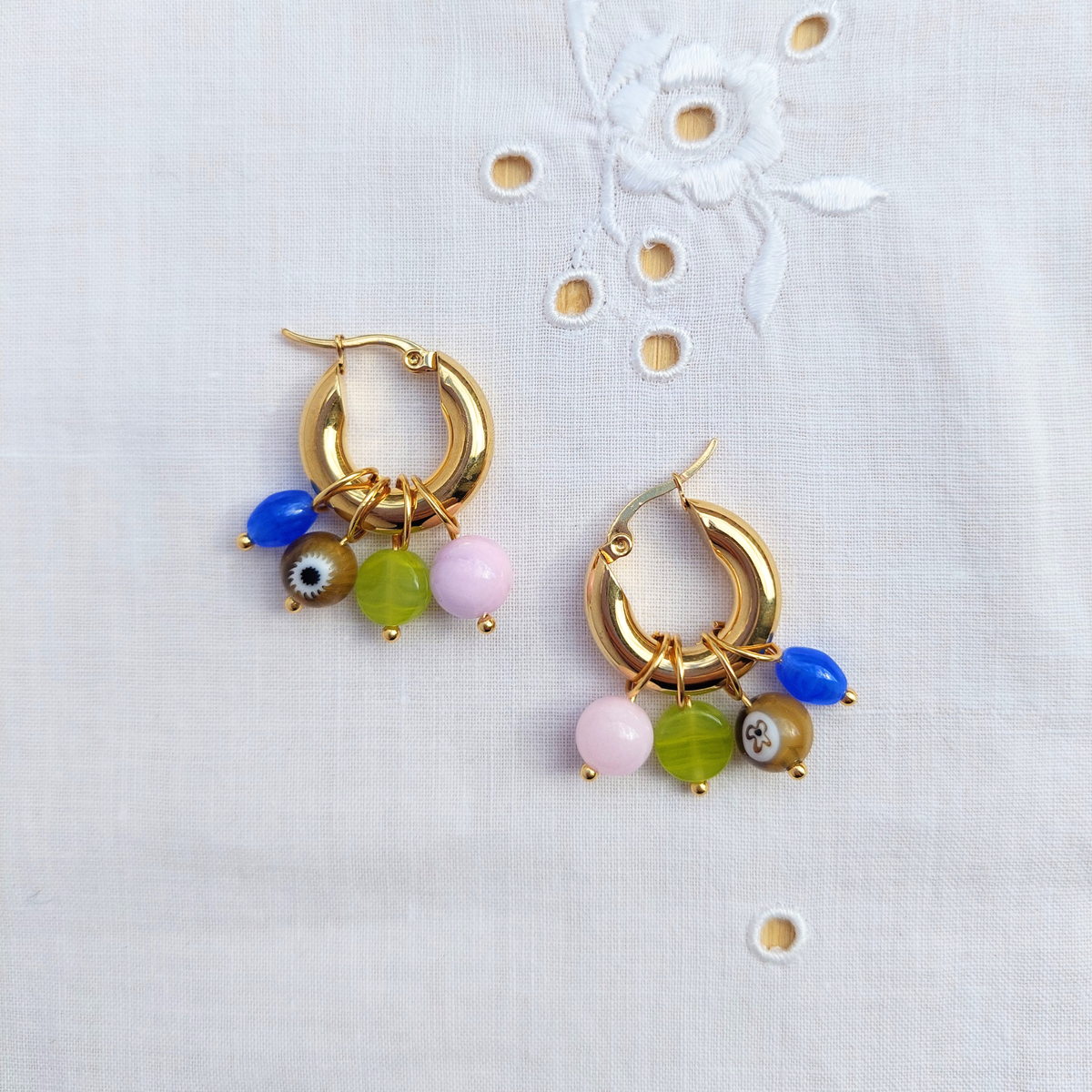 A pair of gold hoop earrings with dangling charm beads in blue, pink, green and brown, displayed on a white embroidered fabric background.